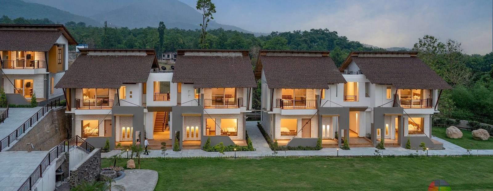 A row of luxurious villas with lit windows, surrounded by lush greenery and mountains in the distance.