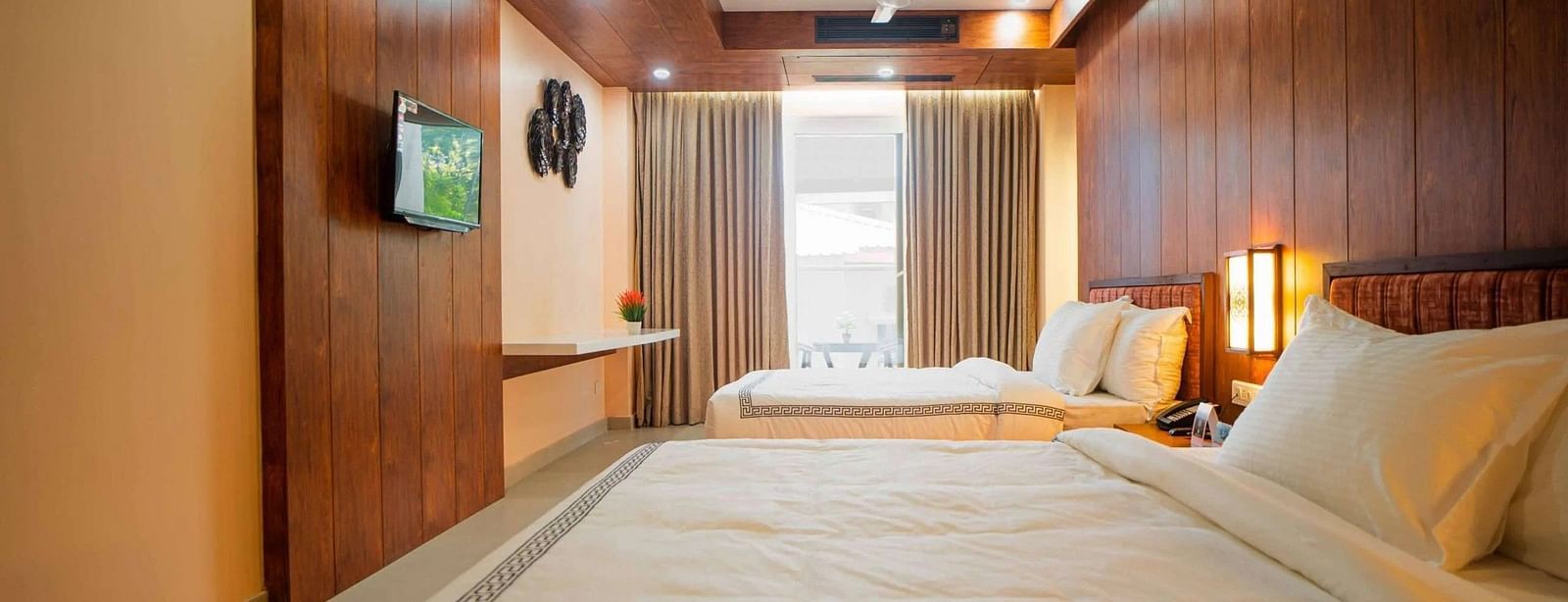 A hotel room at Mansha Palace features two beds, a flat-screen TV, a desk, and curtains partially drawn over a window.