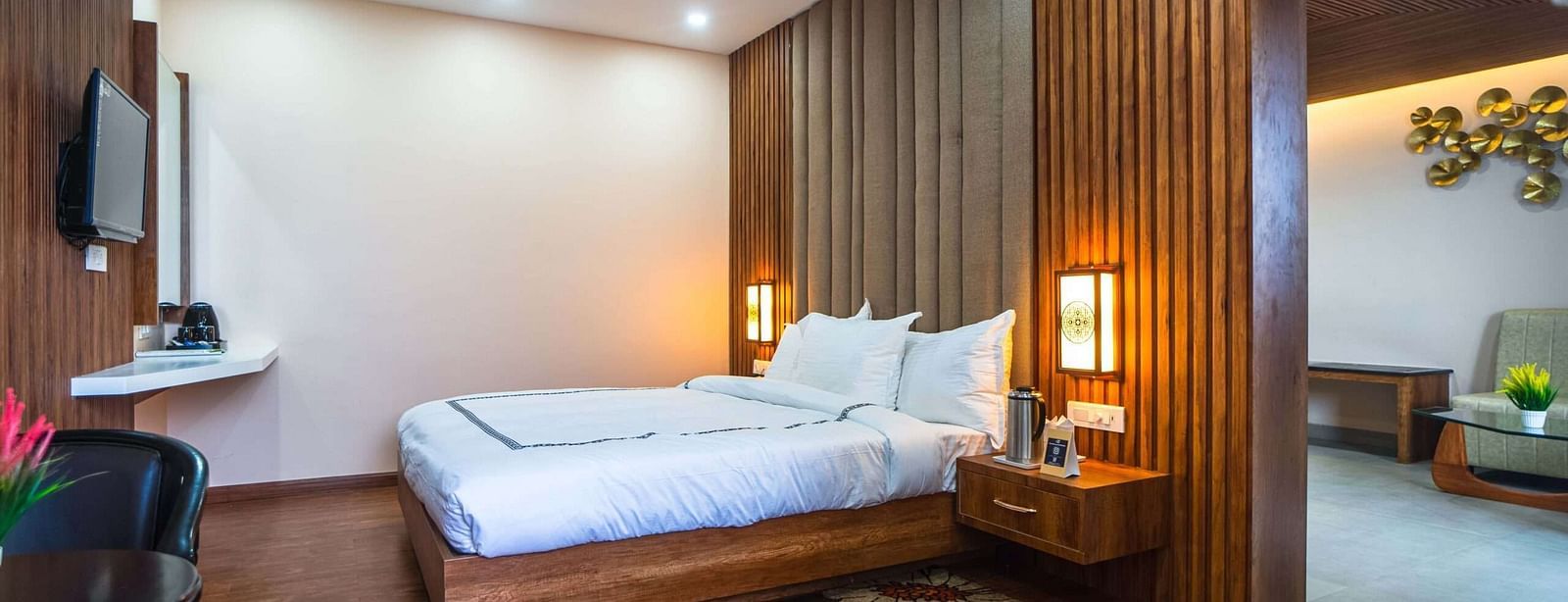A hotel room at Mansha Palace features a bed, nightstands with lamps, a flat-screen TV, a ceiling fan, and a partial view into a seating area.
