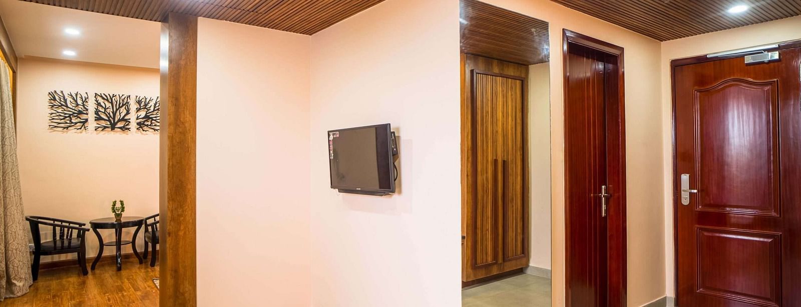 A hotel room entryway shows a flat-screen TV, a full-length mirror, a closed wooden wardrobe, and an exit door. An adjacent seating area is visible.