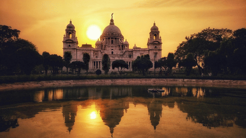 A view of Victoria Memorial, Kolkata, reflected in the water during a golden sunset with trees on either side.
