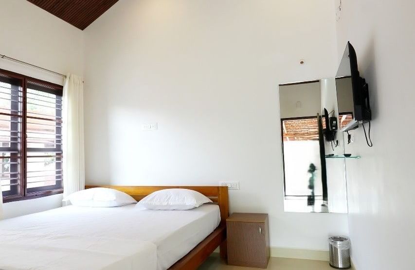 A spacious deluxe room at Bhuthanakadu Retreat, featuring a queen-sized bed with crisp white linens, a wooden headboard, and a high, pitched wooden ceiling with a fan.