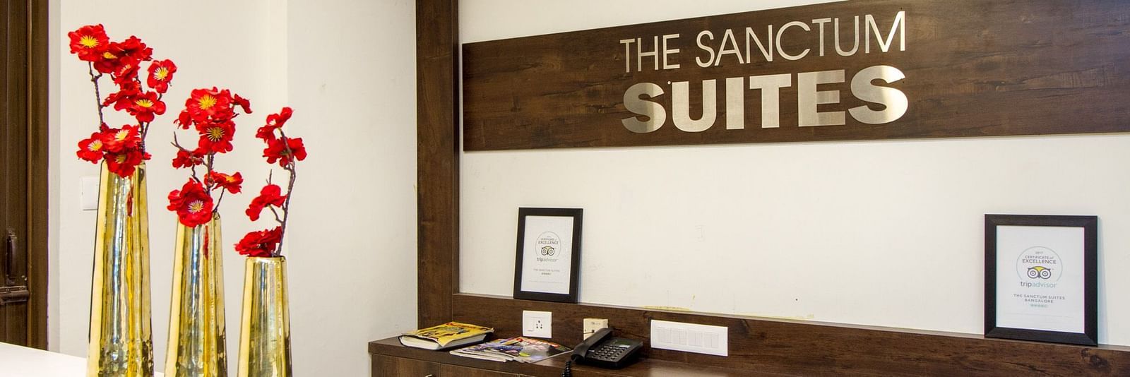 A reception area presenting 'The Sanctum Suites' sign and red flowers placed on a desk at Sanctum Suites Whitefield, Bangalore.