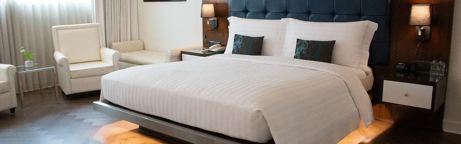 Luxurious hotel suite bedroom with illuminated platform bed, tufted headboard, vibrant artwork, cream armchairs, and warm ambient lighting throughout at Night Hotel Bangkok.