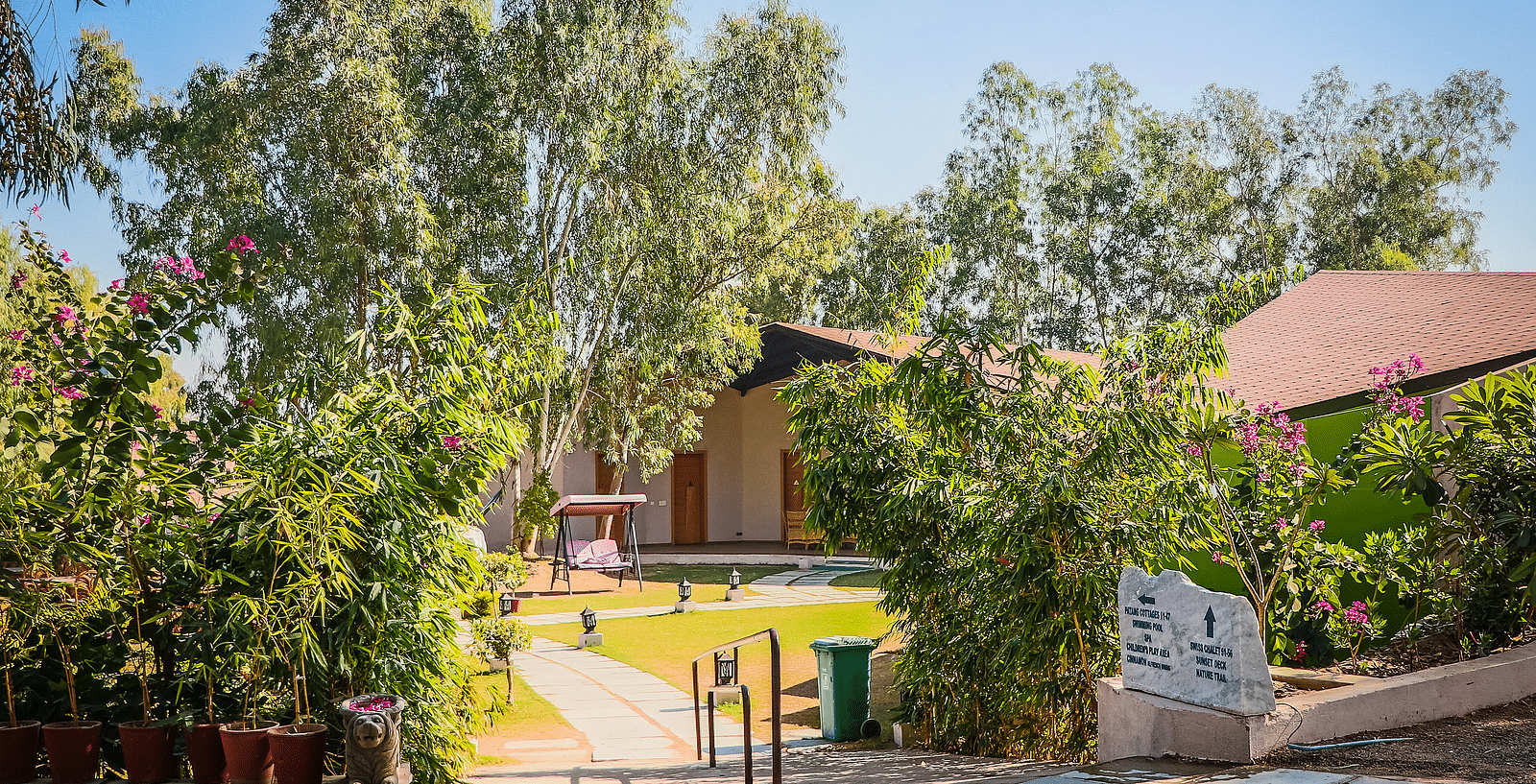 A pathway leads to a building with a sloping roof, surrounded by lush greenery and flowers.- Bamboo Saa Resort & Spa, Udaipur