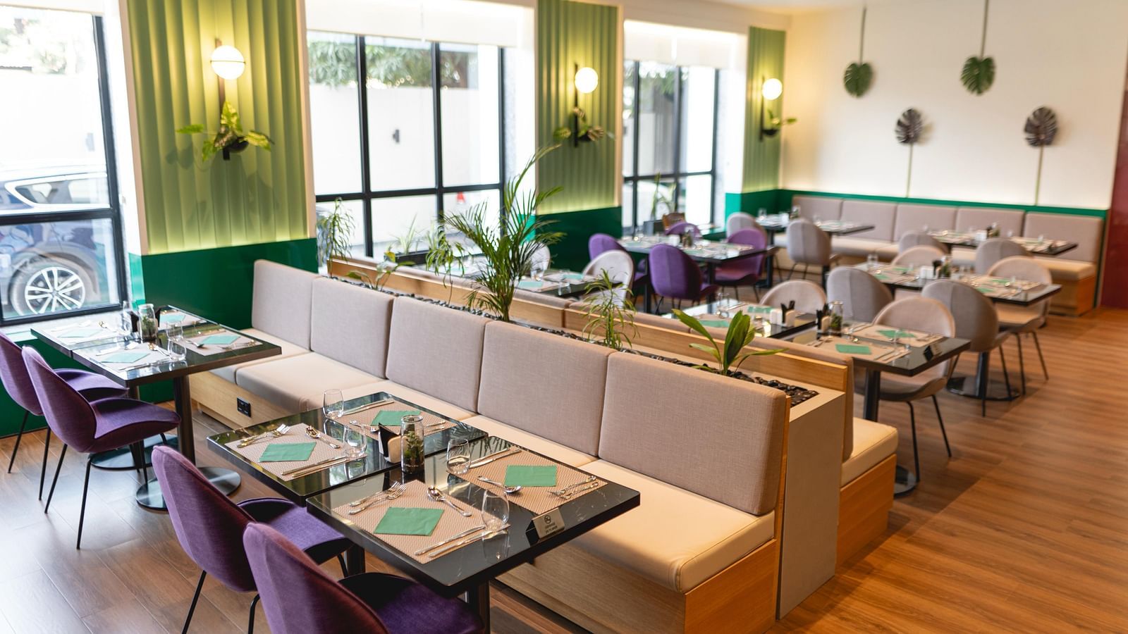 Bright, modern Meli-Melo restaurant interior with accents, large windows, potted plants, and neatly set tables with chairs and beige booth seating. Natural light fills the stylish dining area