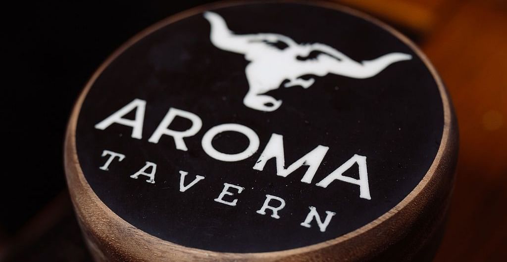 A branded drink coaster featuring the 'Aroma Tavern' logo with a bull's head design, placed on a wooden surface at Shaver Lake Village Hotel.