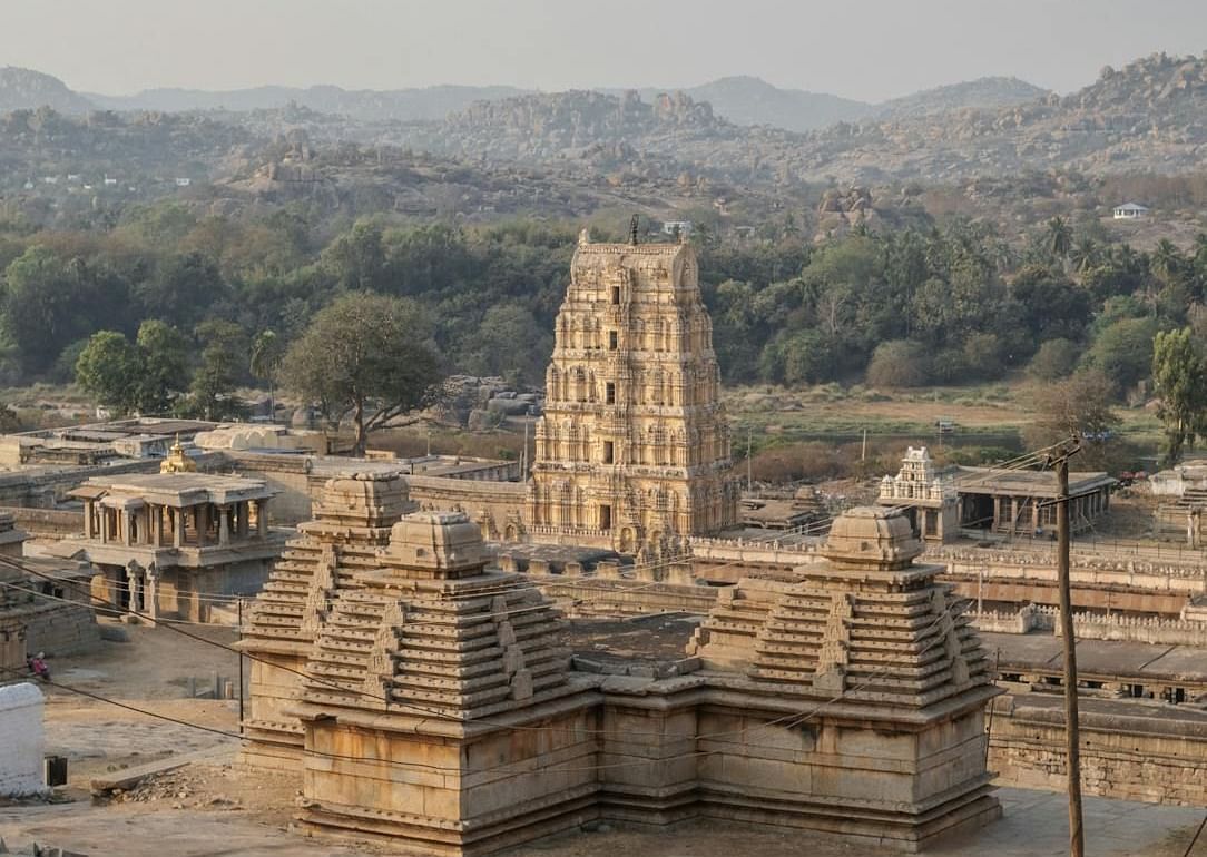 An ancient temple with a tall tower stands amidst a rural landscape with hills in the background.