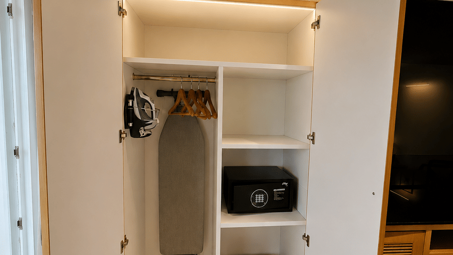 An open wooden wardrobe showing shelves, an ironing board, and a small safe at BluSalzz Collection - Casa Onze, Arpora.