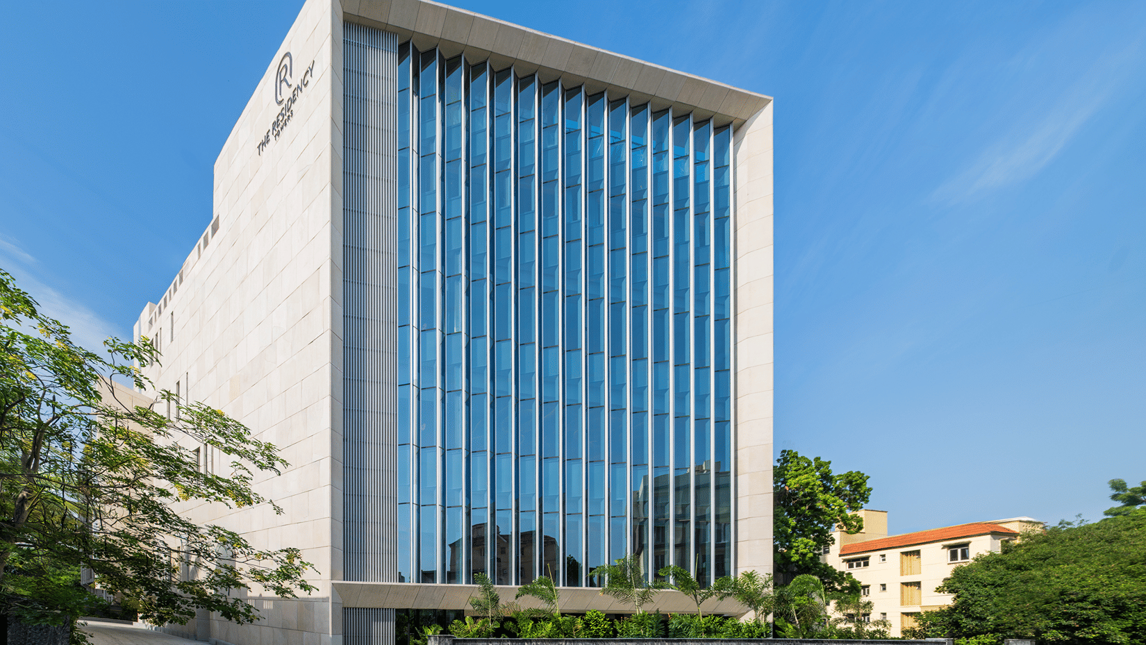 Hotel facade in daylight with white stone cladding, vertical blue glass fins, a stone boundary wall and trees by the driveway at The Residency Towers Chennai.
