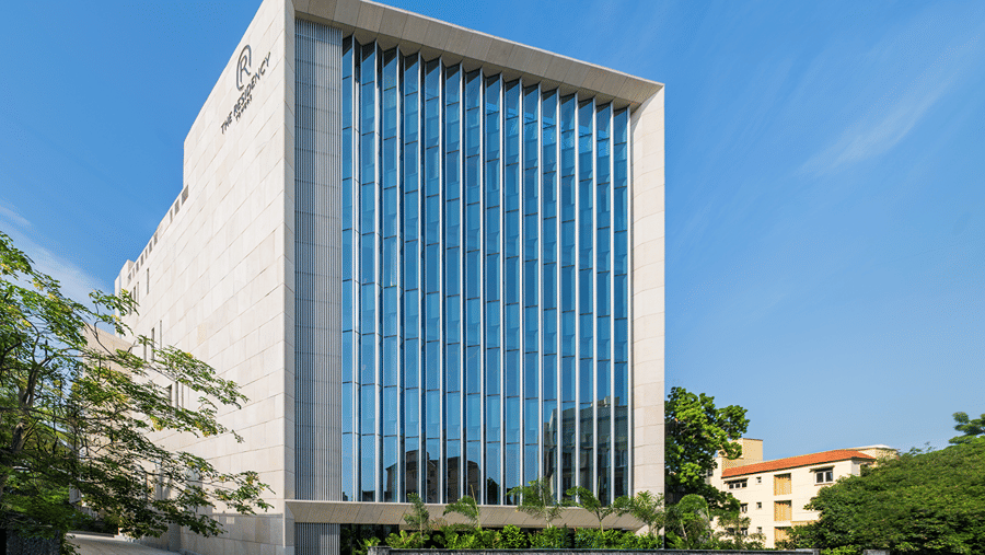 Hotel facade in daylight with white stone cladding, vertical blue glass fins, a stone boundary wall and trees by the driveway at The Residency Towers Chennai.