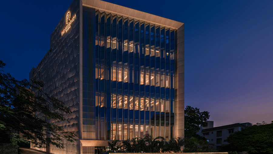 Hotel facade at dusk with illuminated vertical glass fins, rooftop signage and a lit boundary wall along the road at The Residency Towers Chennai.
