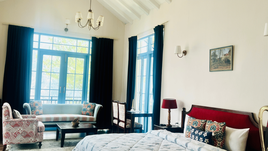 A spacious room at Faraway Cottages, one of the best hotels to stay in Auli, featuring a high vaulted ceiling, a large bed with a red headboard, a seating area with floral armchairs, and large windows framed by dark curtains.