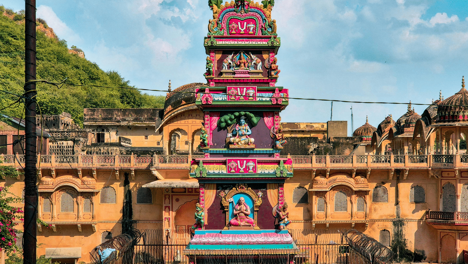  A vertical, brightly painted Hindu shrine or column, with three tiered levels decorated with colorful sculptures of deities and architectural motifs, stands in front of a low, yellow-ochre colored building complex and a green, scrub-covered hill under a v