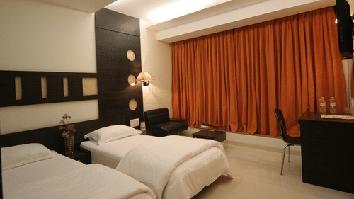 Business Rooms at Pride Biznotel Alkapuri featuring twin beds placed in front of the TV mounted on the wall and a work desk next to the window behind which a sitting area is organised 