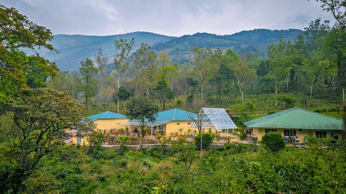 Ibex Resorts, Valparai (Mistly) buildings with green roofs located on a hillside, surrounded by trees, plants, pathways, and mountain ranges in the background under a cloudy sky.