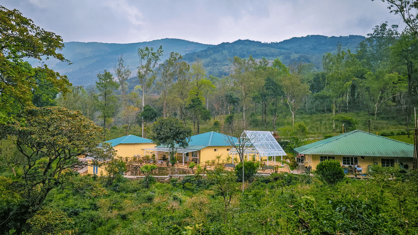 Ibex Resorts, Valparai (Mistly) buildings with green roofs located on a hillside, surrounded by trees, plants, pathways, and mountain ranges in the background under a cloudy sky.