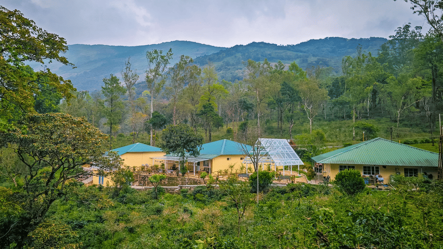 Ibex Resorts, Valparai (Mistly) buildings with green roofs located on a hillside, surrounded by trees, plants, pathways, and mountain ranges in the background under a cloudy sky.