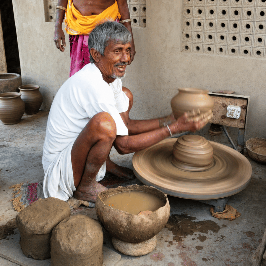 An artisan shaping a clay pot on a traditional potter's wheel while a woman stands nearby, surrounded by handcrafted earthenware pots outdoors.