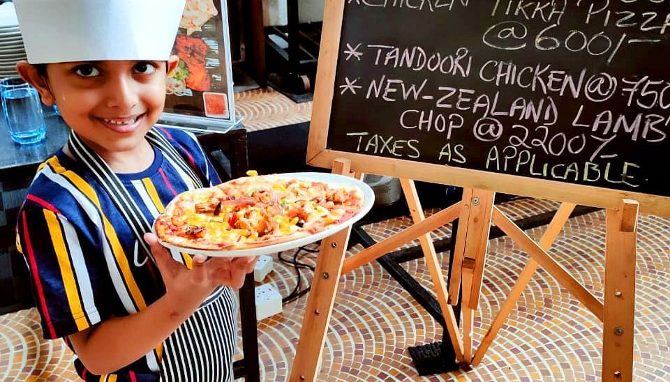 a child showing a pizza after making it in front of a signboard - Caravela Beach Resort Goa
