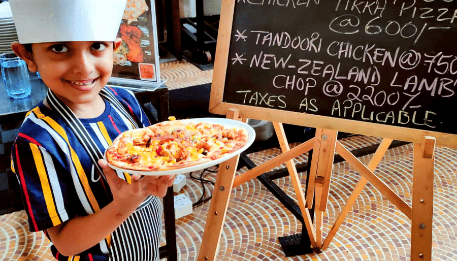 a child showing a pizza in front of a signboard after making it - Caravela Beach Resort Goa.