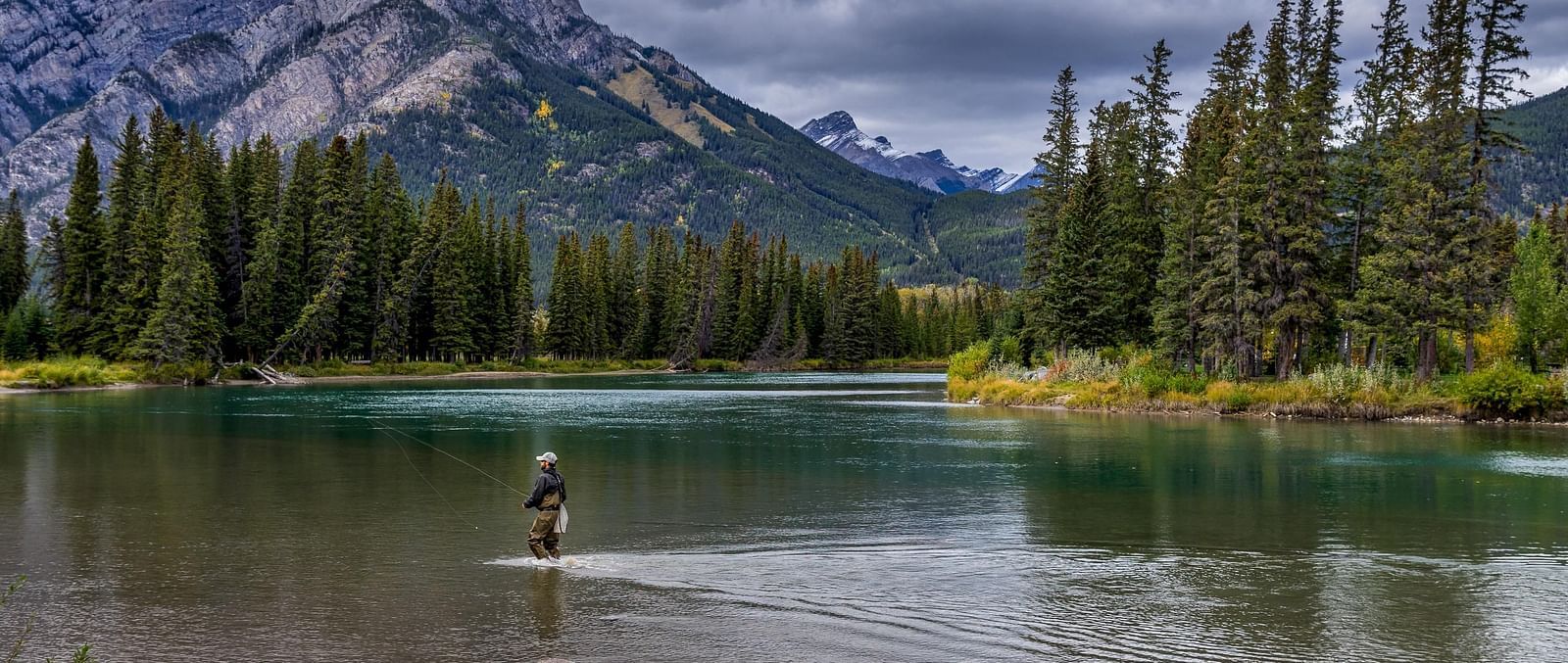 A lone person fishing in a river surrounded by greenery and a mountain.