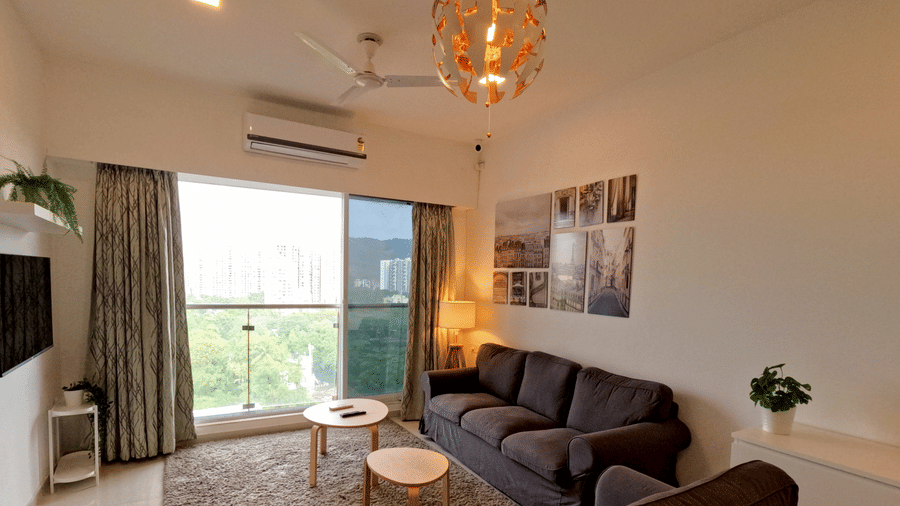 A full view of a living room with a sofa and glass windows at Sahil Exotica by Maxxvalue Apartemnt