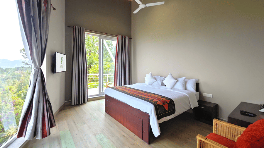 Bedroom interior in Hornbill at Ibex Resorts, Coonoor (Tapas). A bed sits against the wall with pillows and a runner. Windows with curtains flank the bed, showing a view outside. A ceiling fan hangs above, and an armchair sits in the foreground.