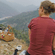 A woman in a red top sitting on a rocky ledge with a trekking pole, looking out over misty mountains.