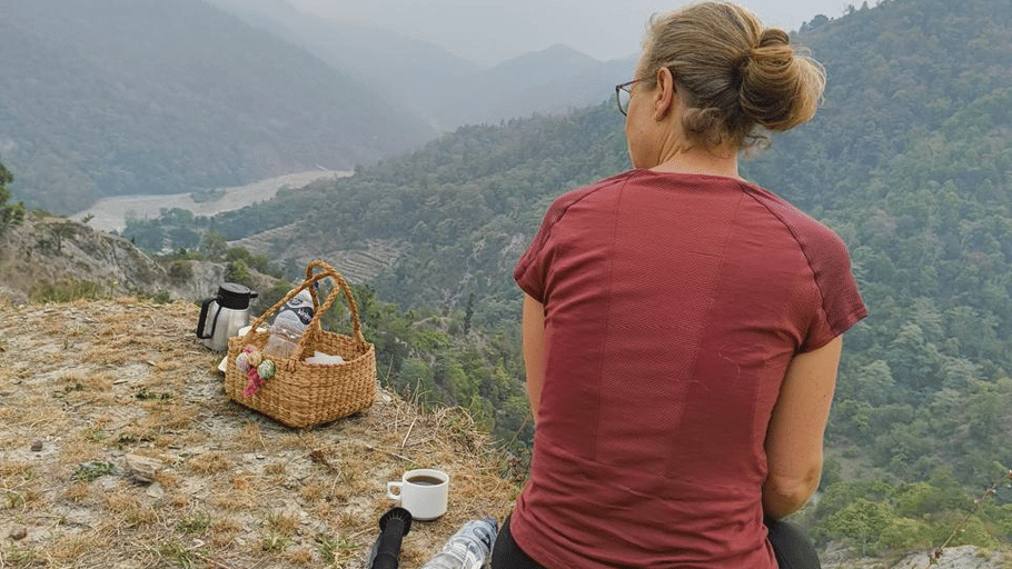 A woman in a red top sitting on a rocky ledge with a trekking pole, looking out over misty mountains.