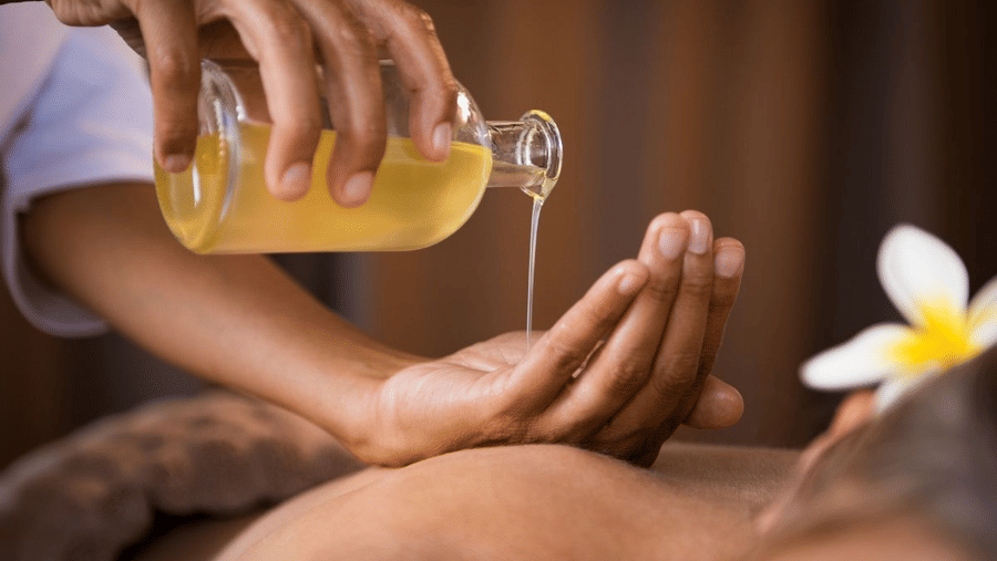 Close-up of a person pouring warm massage oil into their palm during a spa therapy session, with a flower placed beside the client.