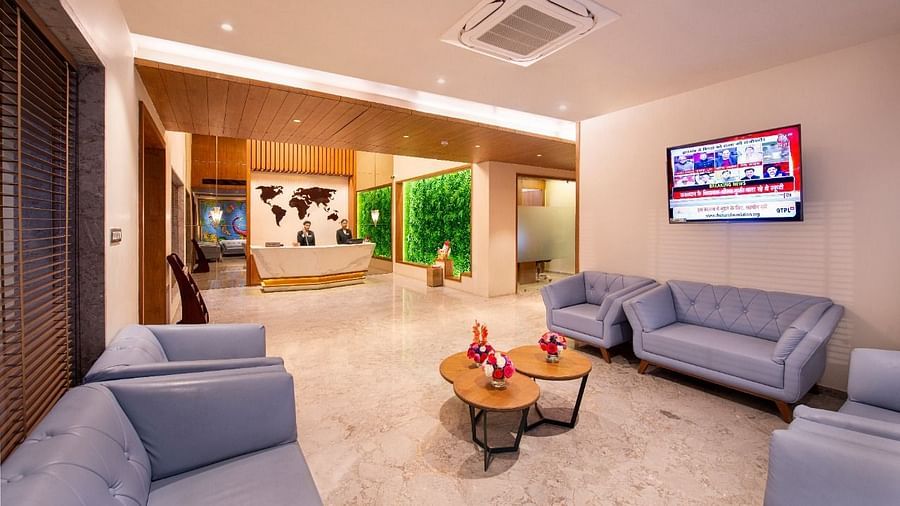 A comfortable hotel lobby lounge area with plush light blue sofas and armchairs arranged around small round tables with floral decorations, a television screen on the wall, and a view towards the reception desk with a world map backdrop and green wall feat