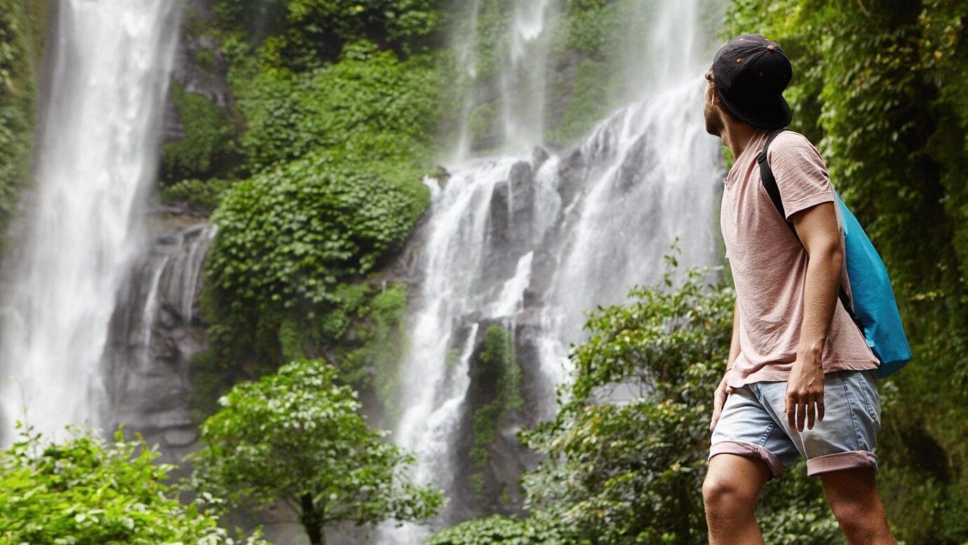 An image of a man looking at the waterfall which is surrounded by trees amidst the hills
