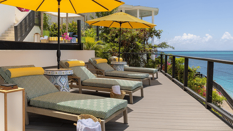 A rooftop deck with lounge chairs and yellow umbrellas overlooks a calm ocean under a clear sky.