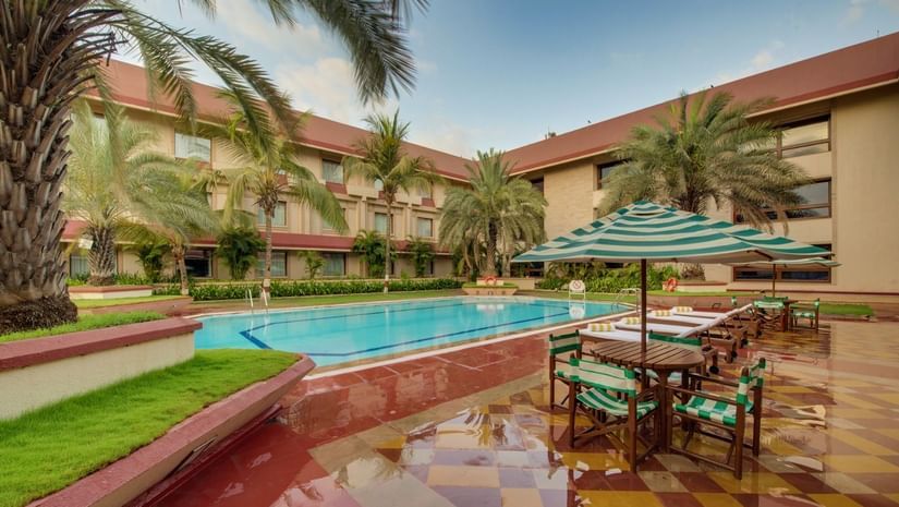 Poolside area at The Ummed Ahmedabad featuring outdoor seating with umbrellas, palm trees, and surrounding hotel buildings under a clear sky.