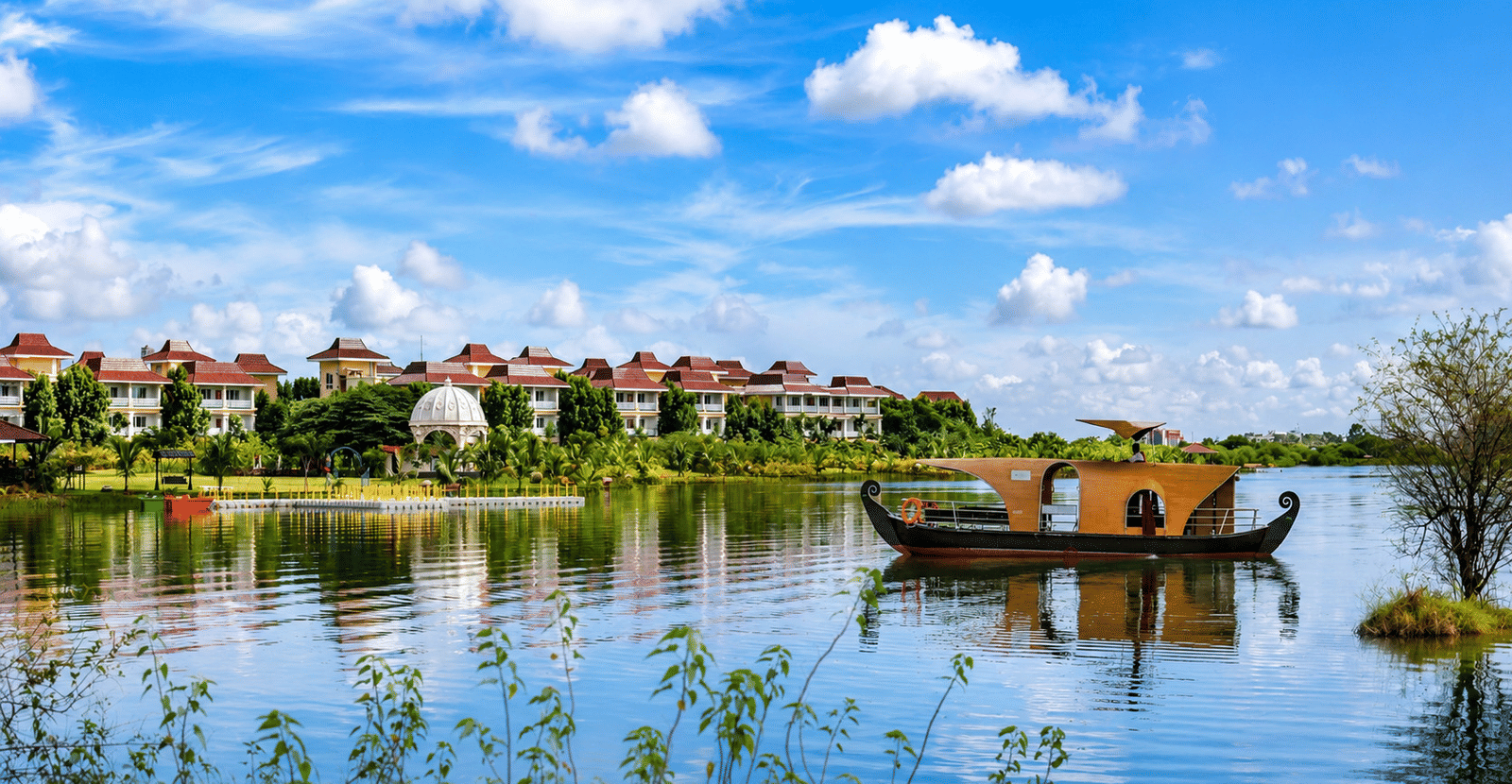 A covered wooden boat sails across a lake in front of the MAYFAIR Lake Resort Raipur under a clear sky.