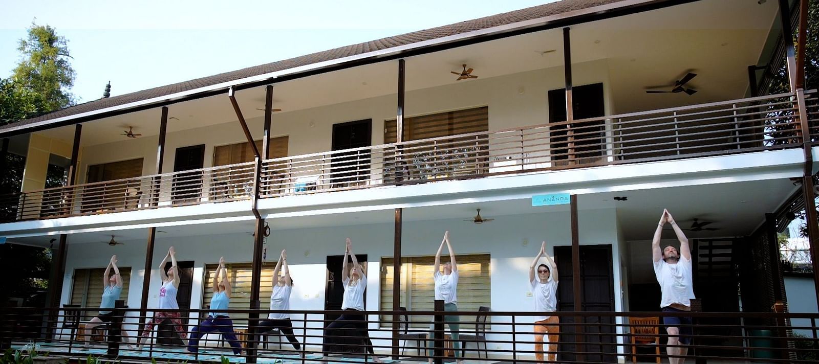 A group of people practice yoga poses on a balcony overlooking a garden, with a multi-story building in the background.
