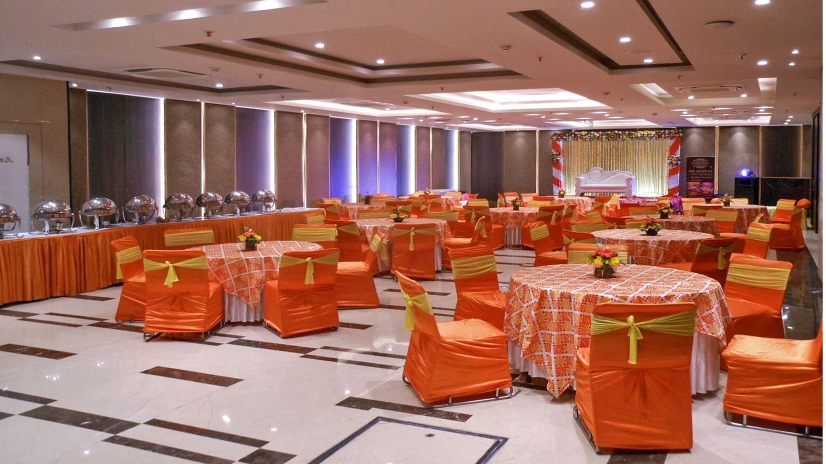 A picture of a banquet hall with decorated round tables and chairs, a buffet section on one side, and a stage at the front featuring a white sofa.