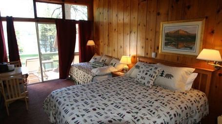A cosy cabin room with two beds and patterned comforters, warm wood paneling, a framed picture and  glass door leading to a balcony. - Kit Carson Lodge & Restaurant