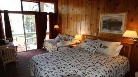 A cosy cabin room with two beds and patterned comforters, warm wood paneling, a framed picture and  glass door leading to a balcony. - Kit Carson Lodge & Restaurant