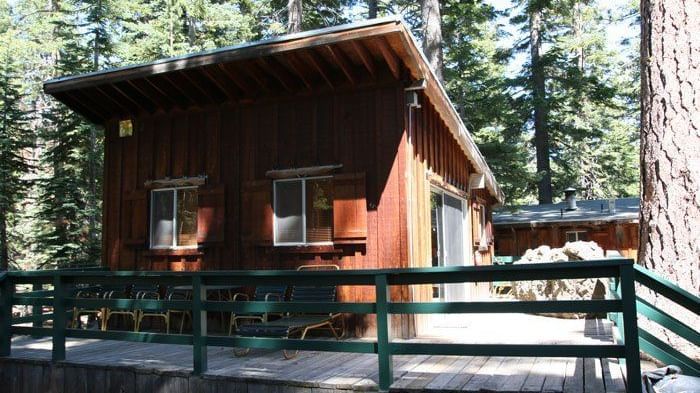 A rustic cabin with a steeply slanted roof, small windows, and weathered wood siding sits nestled among tall pine trees. -  Kit Carson Lodge