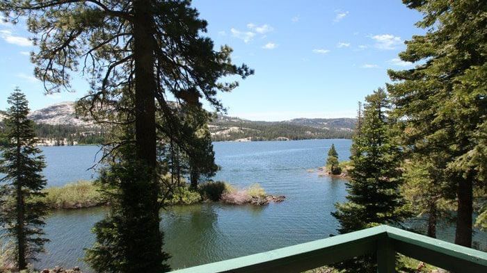 A scenic lake view framed by pine trees and a green railing, with distant mountains under a blue sky.- Kit Carson Lodge & Restaurant 