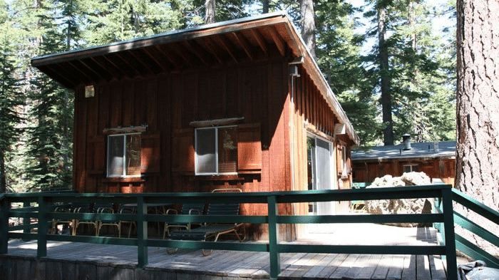 A rustic cabin with a steeply slanted roof, small windows, and weathered wood siding sits nestled among tall pine trees. -  Kit Carson Lodge
