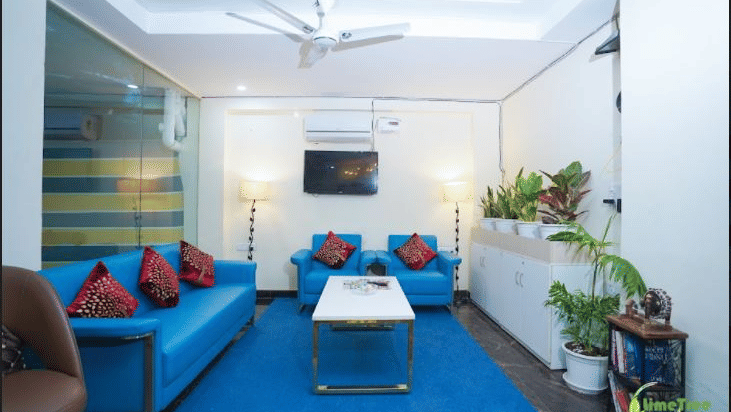 A contemporary waiting lounge featuring bright blue sofas, a central coffee table, a blue rug, and potted indoor plants.