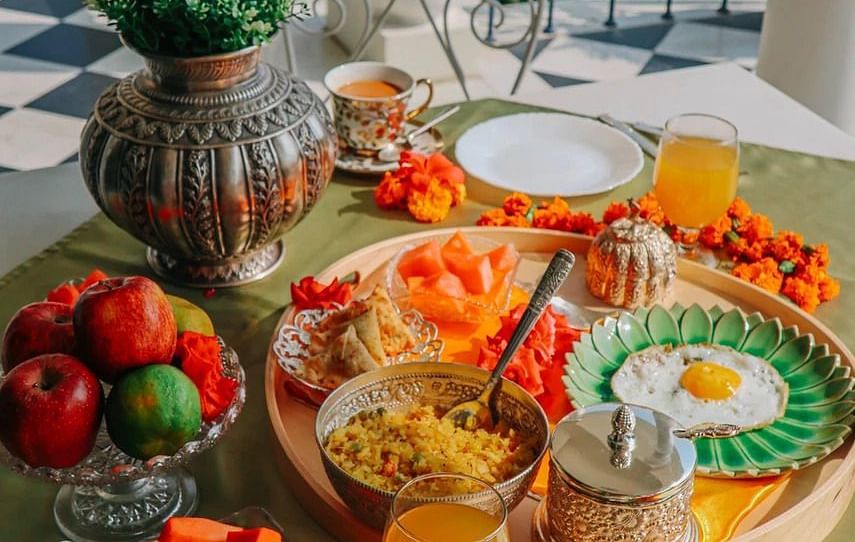 A breakfast spread laid out on a table on a balcony, featuring fruits, eggs, drinks, and other dishes, with a view of blue railings and greenery | Dileep Kothi, Jaipur