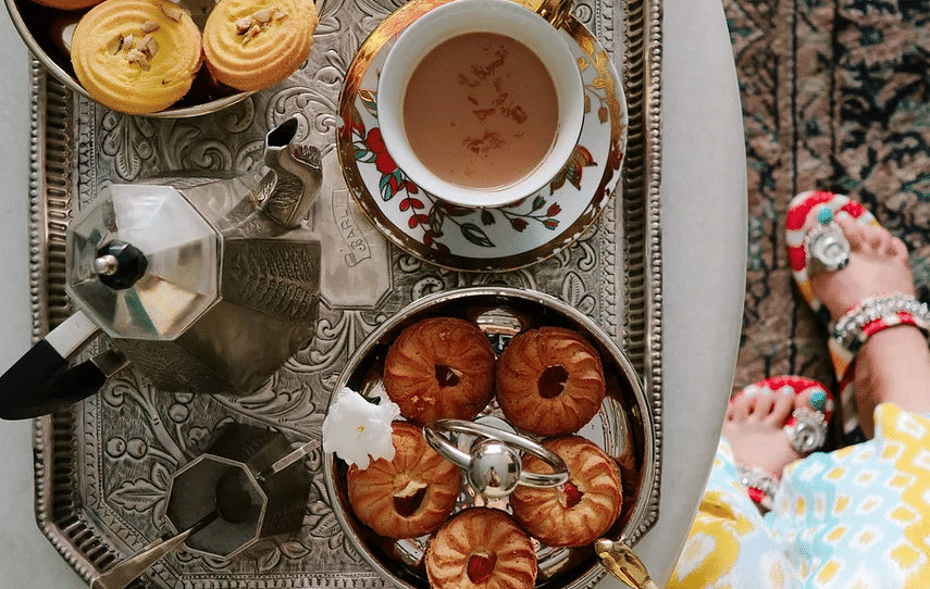 A flat lay of a silver tray with coffee, pastries, and a yellow flower on a white table, with a person's patterned dress and feet visible at the edge | Dileep Kothi, Jaipur