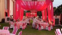 A lush green lawn set up for an evening event with pink-draped tables, matching chairs, and a large decorative canopy at Golden Tulip Kukas, Jaipur.