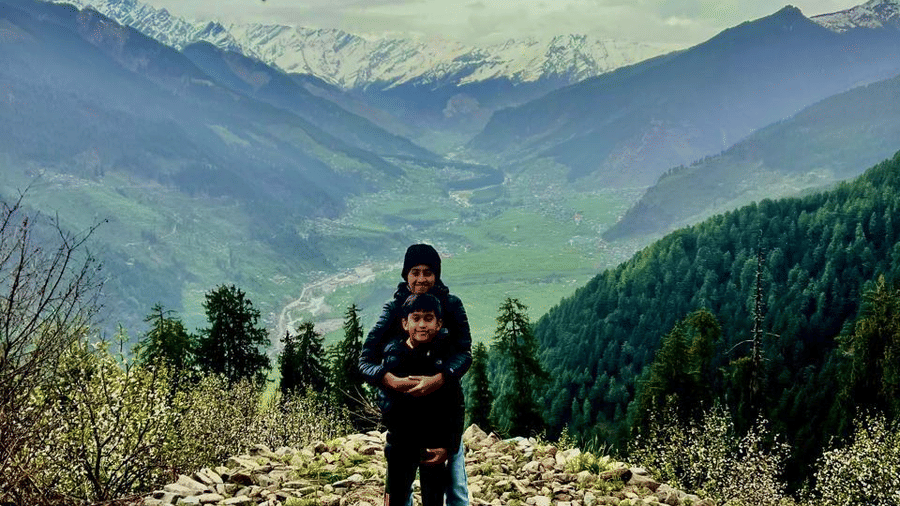 2 kids happily posing for a picture on a rocky terrain with a beautiful background featuring the mountains under a cloudy sky at Amara Upepo - The Sky Village, Manali.