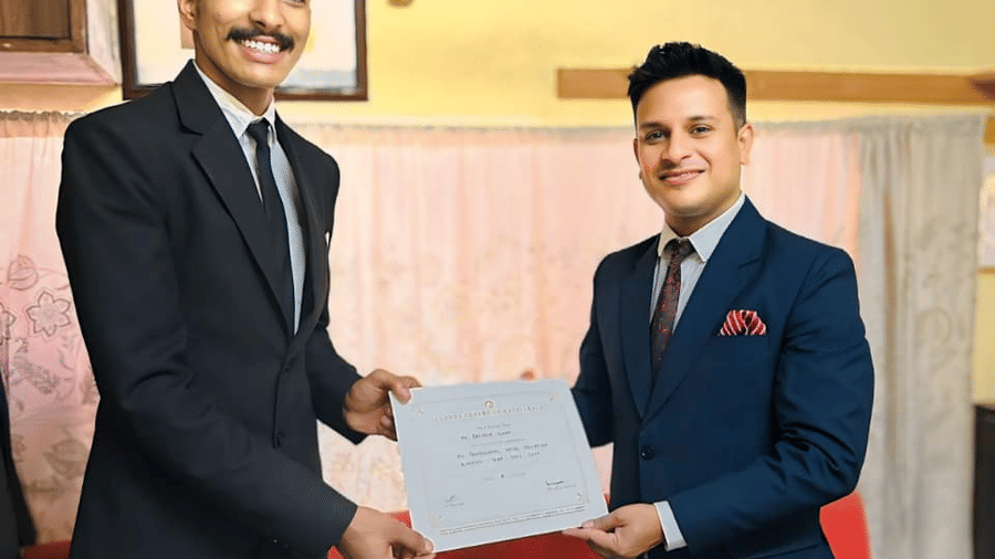 A person in a suit receives a certificate from another individual inside a room at Clarks Group of Hotels. They stand next to a sofa.