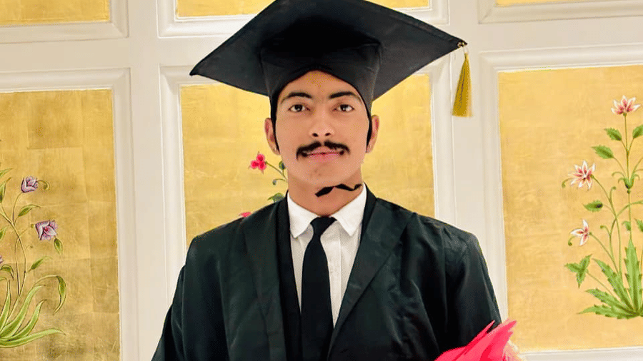 A person wearing a graduation gown and cap stands indoors. The person holds a bouquet of flowers and a box.